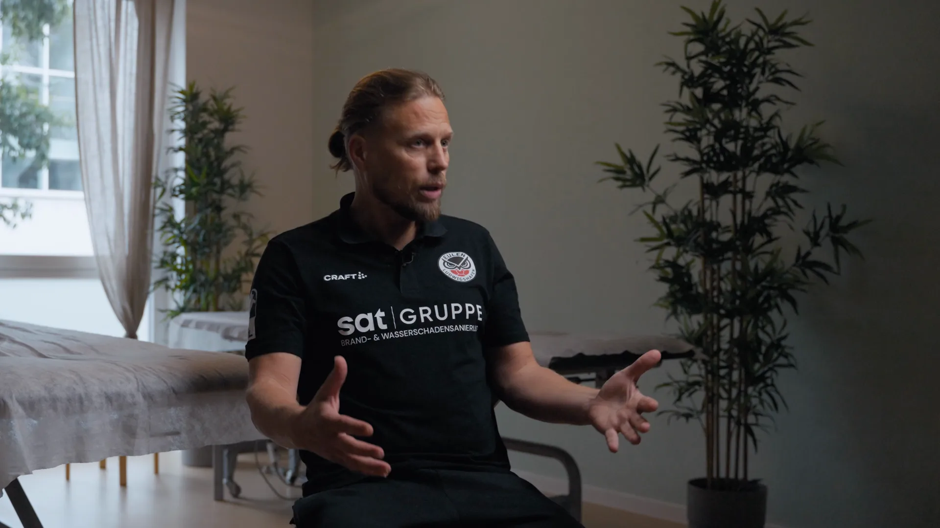Ein Mann im Polo-Shirt erzählt im Interview, im Hintergrund Pflanzen und ein helles Fenster.