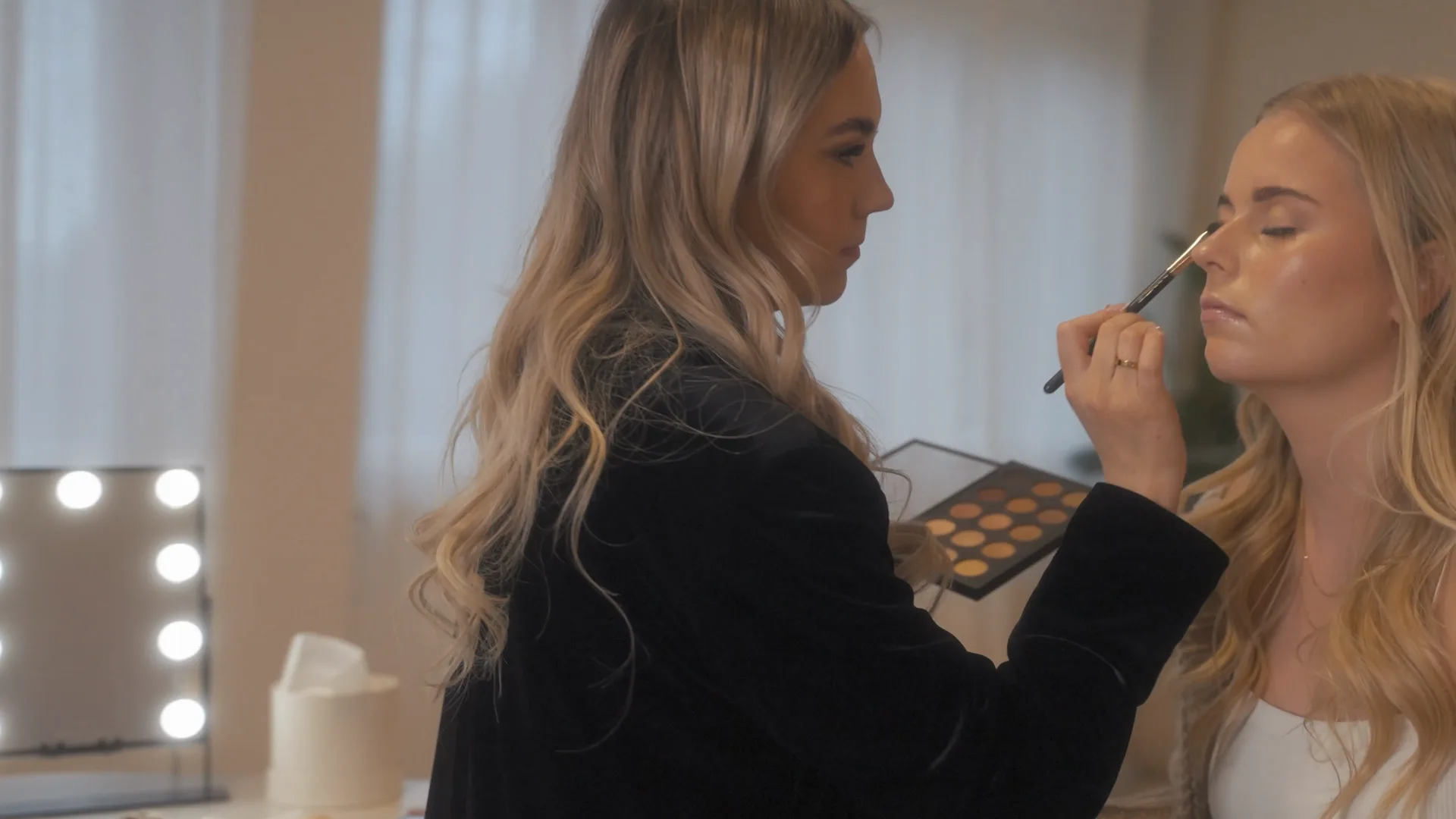 Eine Make-up-Artistin schminkt eine junge Frau vor einem beleuchteten Spiegel.