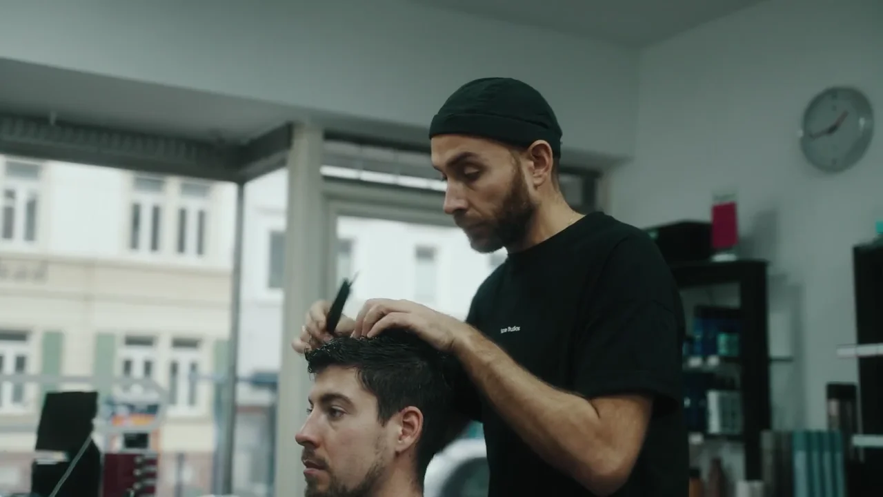 Ein Friseur mit Mütze schneidet einem Kunden am Fenster die Haare.